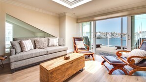 A modern living room with a grey sofa, wooden coffee table, and a wooden armchair with a view of the marina through large glass doors. - Luxurious Channel Islands Harbor Home w/ Boat Dock (Oxnard)