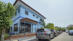 Edifício de dois andares com fachada azul e telhado vermelho. Possui uma área externa coberta com guarda-corpo de metal e uma escadaria que leva à entrada. Vários carros estão estacionados em frente ao prédio.