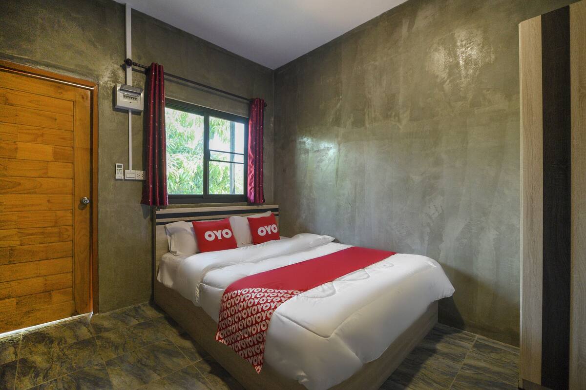 A bedroom with a wooden door, a window with red curtains, a bed with white bedding and red pillows, and a wall-mounted air conditioning unit.