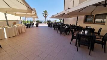 Un espace de restauration en plein air, avec des tables et des chaises, protégé par de grands parasols, et offrant une vue sur la mer en arrière-plan.