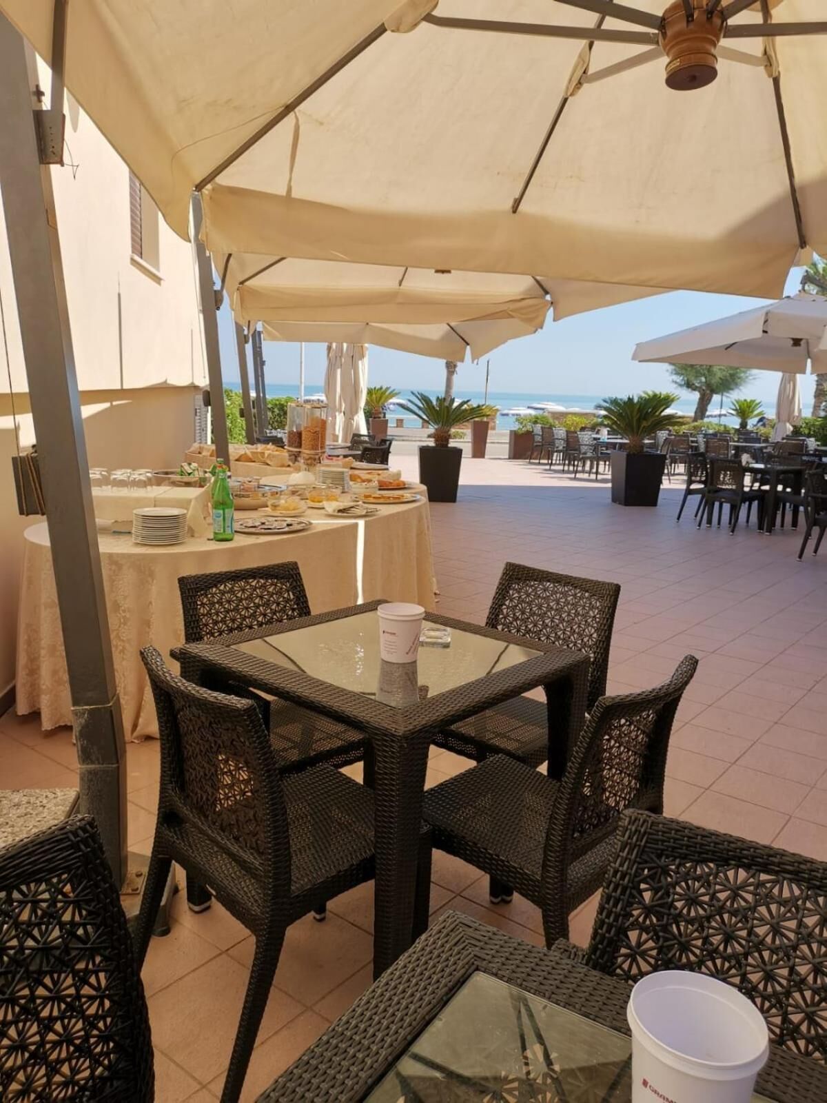 Un área de comedor exterior con sillas de mimbre y una mesa de vidrio, con vista a una playa.