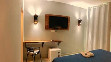 Quarto de hotel com cama, escrivaninha, cadeira, televisão e um pequeno eletrodoméstico branco.