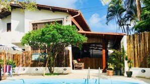 Una piscina con un borde de baldosas, rodeada por una casa de fachada blanca y cerca de madera.