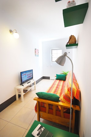 Quarto com cama, escrivaninha, luminária, televisão e janela.