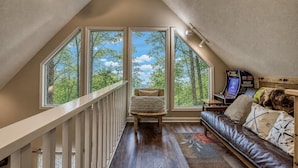 A loft bedroom with a sloped ceiling, large windows, a wooden staircase, a leather sofa, and a gaming console.
