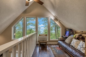 A loft bedroom with a sloped ceiling, large windows, a wooden staircase, a leather sofa, and a gaming console. - Moonshine Chalet, Hot Tub, Pool Access, WIFI,  $560 FREE tickets each paid day! (Gatlinburg)