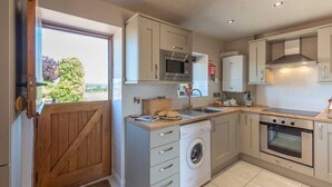A compact kitchen with a built-in oven, microwave, and washing machine, a wooden door, and a view through the window. - The Dinney Holiday Cottages (Bridgnorth)