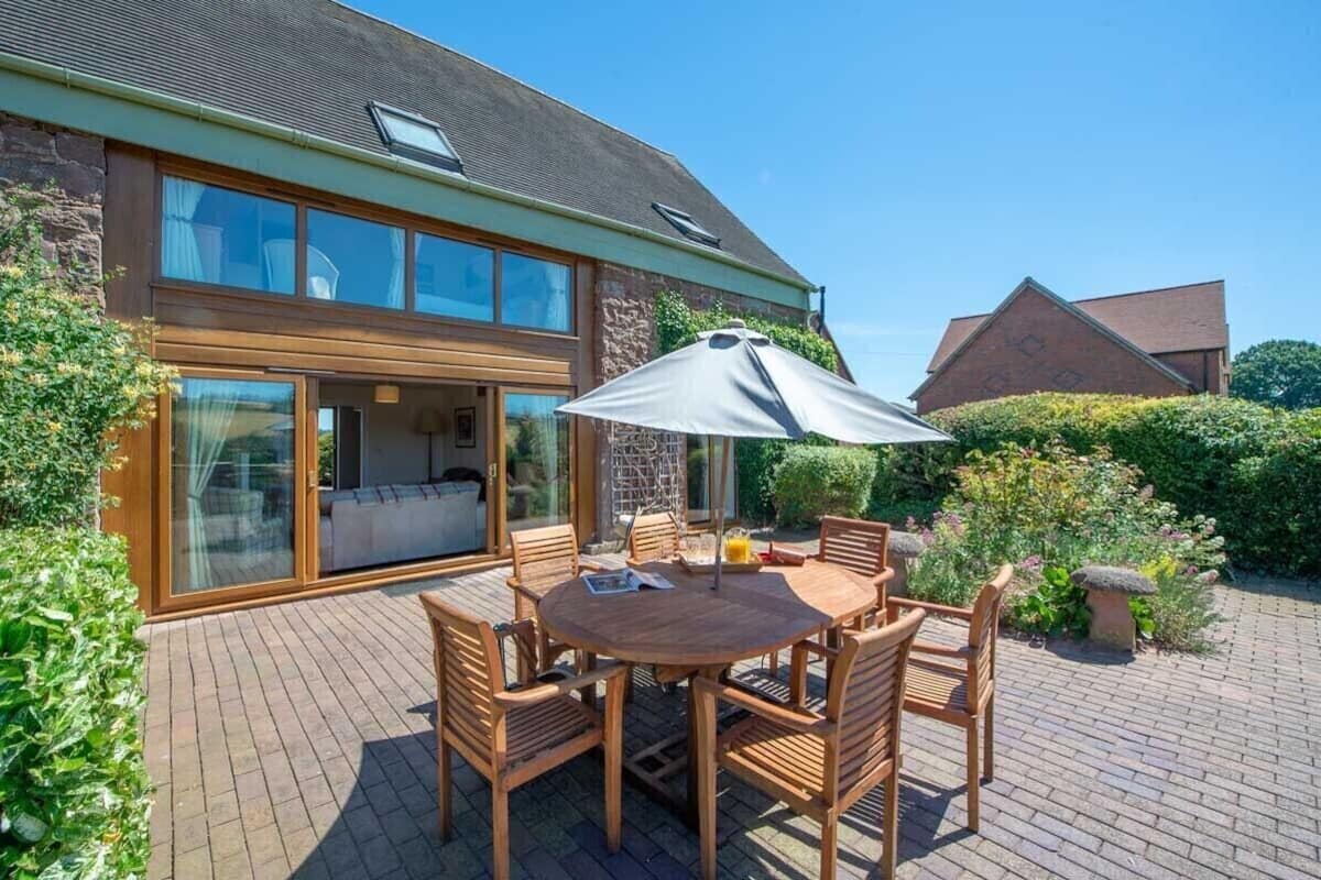 A two-story house with a patio area featuring a round wooden table and chairs, an umbrella, and a brick floor.