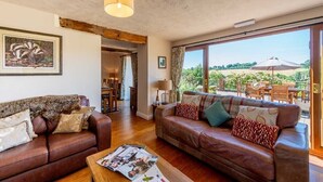 A spacious living room with leather sofas, a wooden coffee table, and a large window offering a view of the outdoors. - The Dinney Holiday Cottages (Bridgnorth)