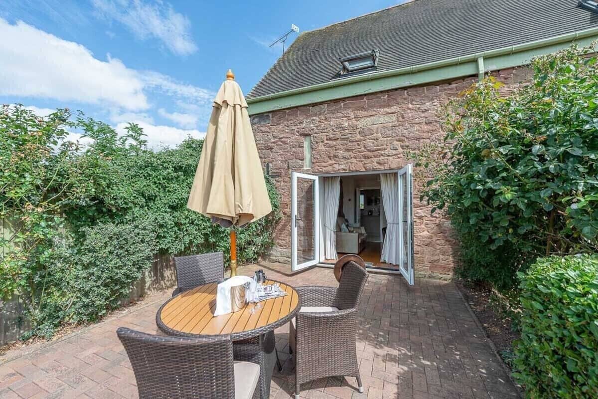 A patio with wicker furniture and a table, a stone building with a green roof, and a garden area with dense foliage.