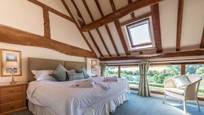 A bedroom with a large bed, wooden beams, a window with a view, a nightstand, and a chair. - The Dinney Holiday Cottages (Bridgnorth)