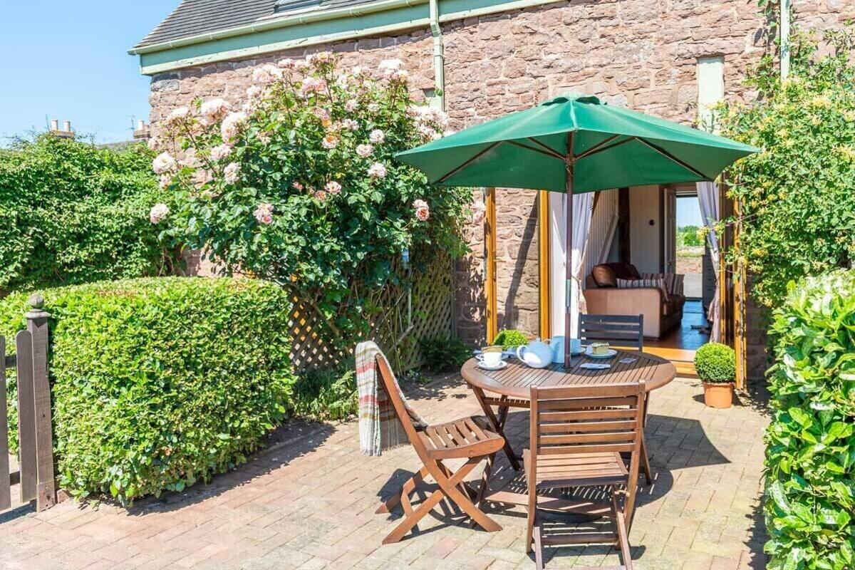 A patio with a wooden table and chairs, a green umbrella, and a view of a stone building with a garden.