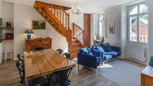 Een ruime woonkamer met een houten trap, een eettafel, blauwe banken en een groot raam.