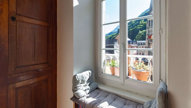 Een kamer met een houten deur, een raam met uitzicht op gebouwen en bergen, en een bank met kussens.