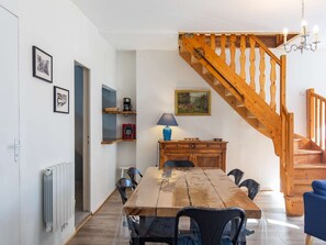 A dining area with a wooden table, chairs, a coffee machine, and a lamp. A staircase with wooden railings leads to an upper level.