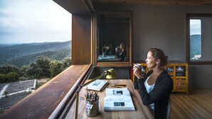 Une pièce dotée d’une grande fenêtre donnant sur un paysage montagneux, d’un bureau avec une lampe et d’une personne tenant une tasse.
