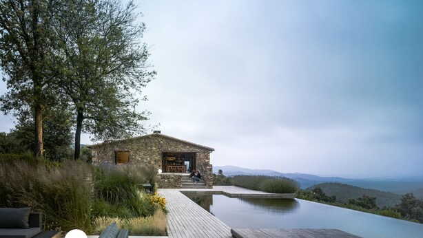 Une maison en pierre dotée d’une terrasse en bois et d’une piscine à débordement, offrant une vue imprenable sur un paysage montagneux.