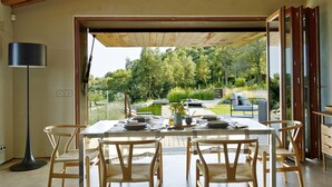 Une salle à manger spacieuse avec des poutres en bois, une table dressée pour quatre personnes et une vue sur un jardin luxuriant, visible à travers de grandes portes vitrées.