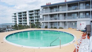 Et poolområde med træterrasse, en pool med metalstige og en bygning med flere altaner i baggrunden.
