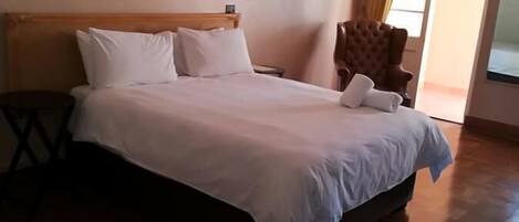 A neatly made bed with white linens in a hotel room with wooden flooring and a large window with curtains.