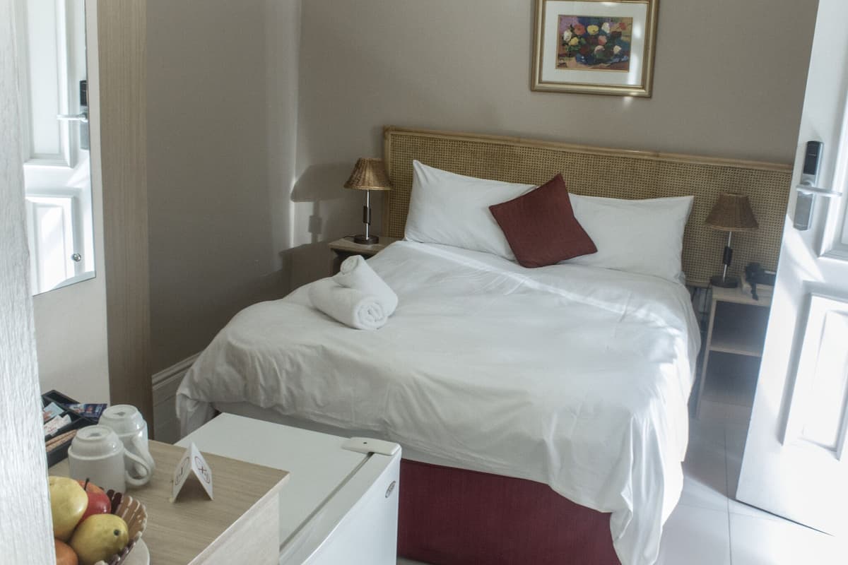 Ein Hotelzimmer mit einem ordentlich bezogenen Bett, einem Nachttisch mit Lampe, einer Obstschale und einem kleinen Kühlschrank.
