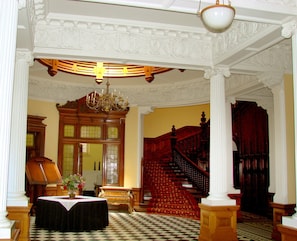 Um salão imponente com piso xadrez, um candelabro, uma escadaria e uma grande escrivaninha de madeira.