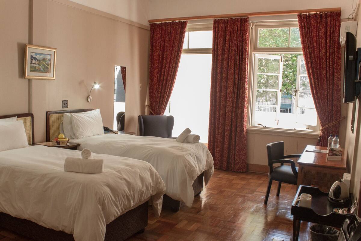 Ein Hotelzimmer mit zwei Betten, einem Schreibtisch mit Stuhl, einem Fenster mit Vorhängen, einem Bild an der Wand und einer Lampe.