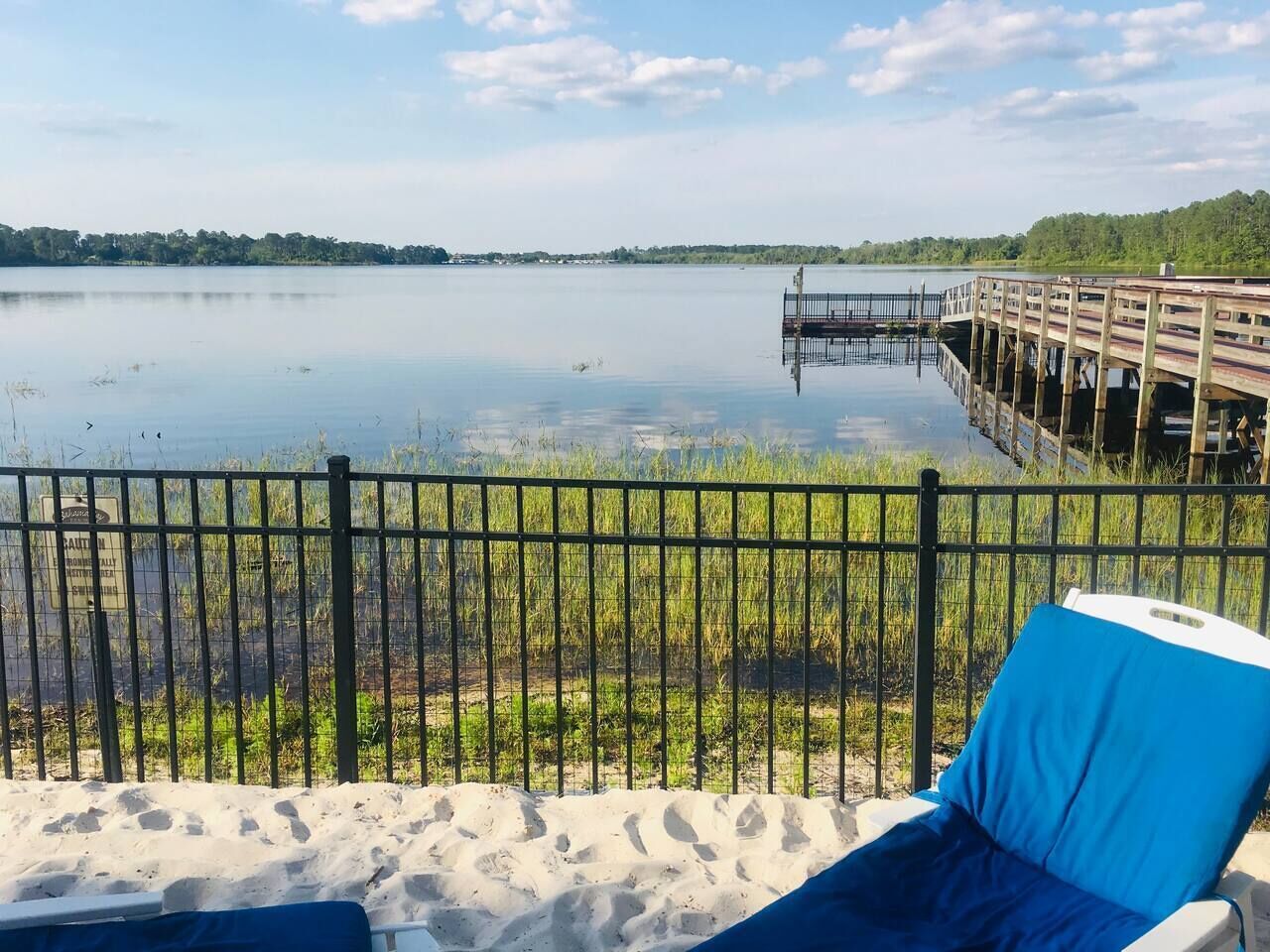Blick auf einen Strand mit einer Holzstege, der sich in ruhiges Wasser erstreckt, einem eingezäunten Sandbereich und einem Liegestuhl mit blauer Polsterung.