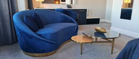 Une chambre d’hôtel moderne avec un canapé bleu, une petite table ronde sur laquelle se trouve un bol, et une vue sur la cuisine et la salle de bain grâce à un miroir.