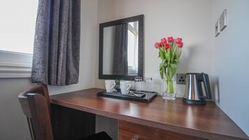 A wooden desk with a mirror, two white cups, a tray, a vase with red flowers, a glass, and a kettle.