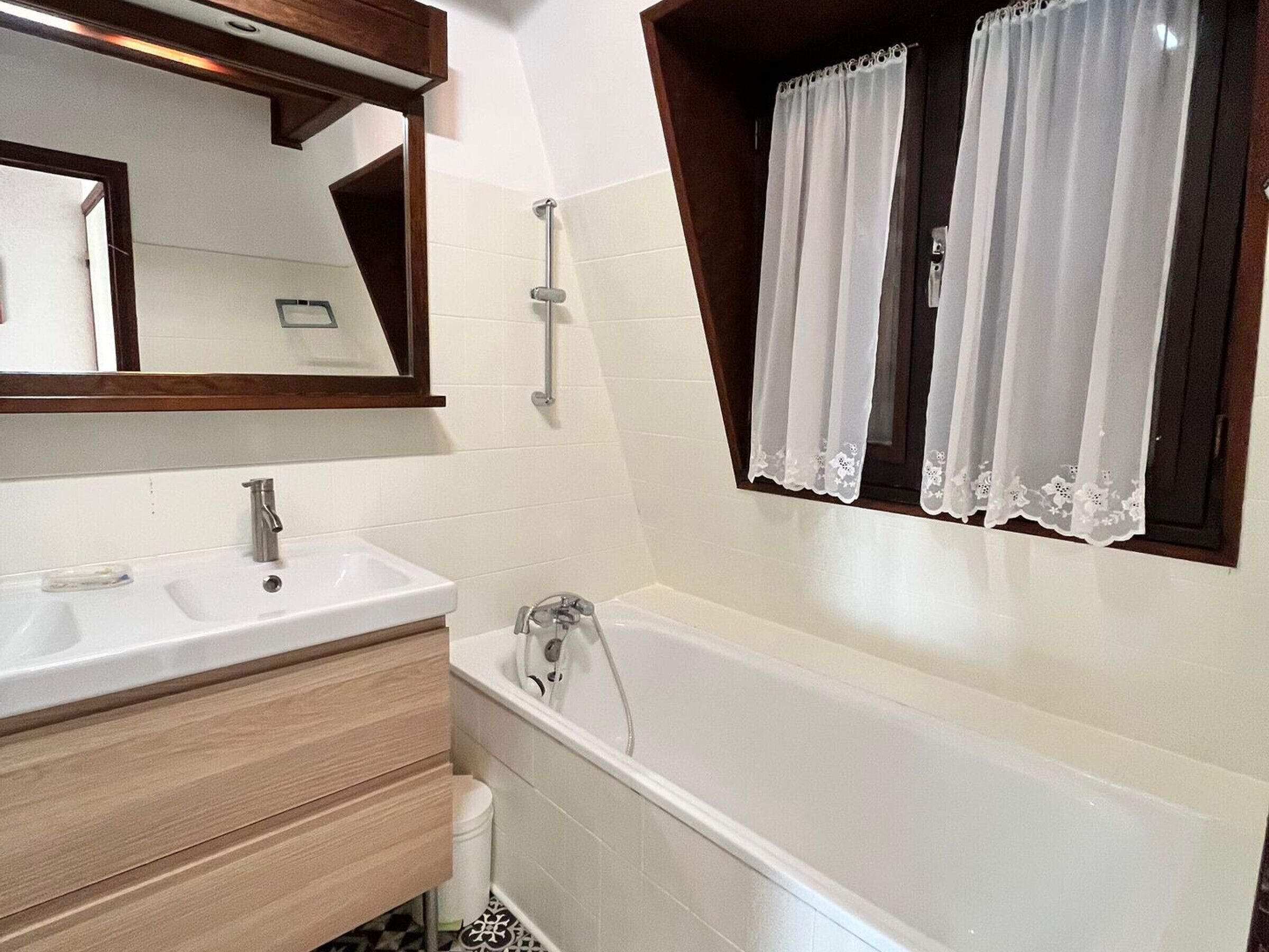 Une salle de bain moderne avec un double vasque, une baignoire et une fenêtre ornée de rideaux en dentelle.