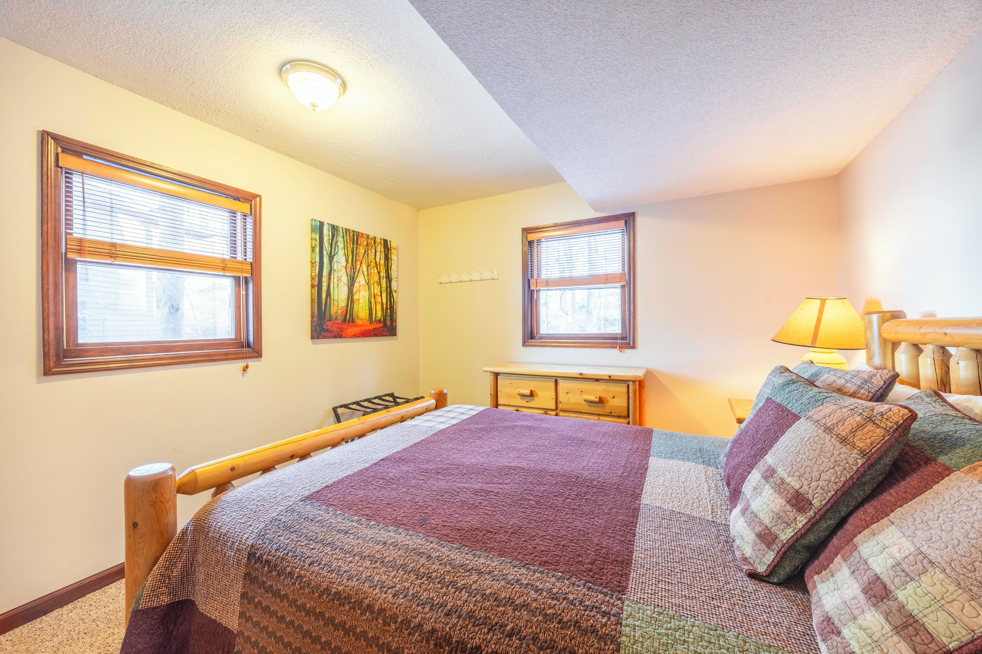 A bedroom with a bed, two windows, a nightstand, a dresser, and a painting on the wall.