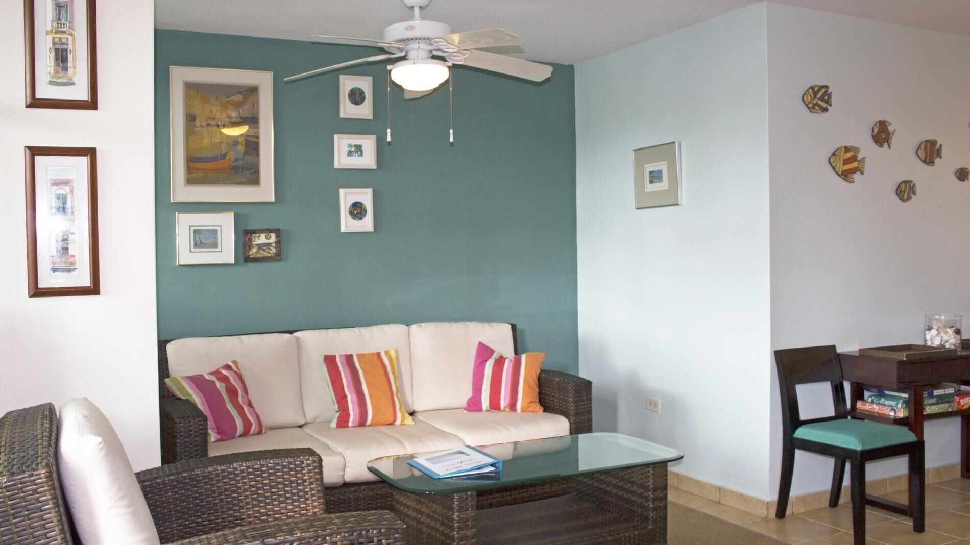 A living room with a sofa, coffee table, and a ceiling fan. There are framed pictures on the walls and a small table with a chair.