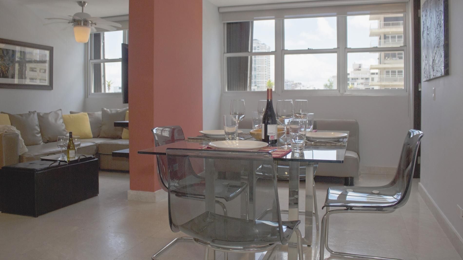 Een moderne eetruimte met een glazen tafel, transparante stoelen en stadszicht door grote ramen.