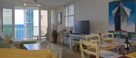 A living room with a dining area, a TV, and a large window overlooking the ocean.