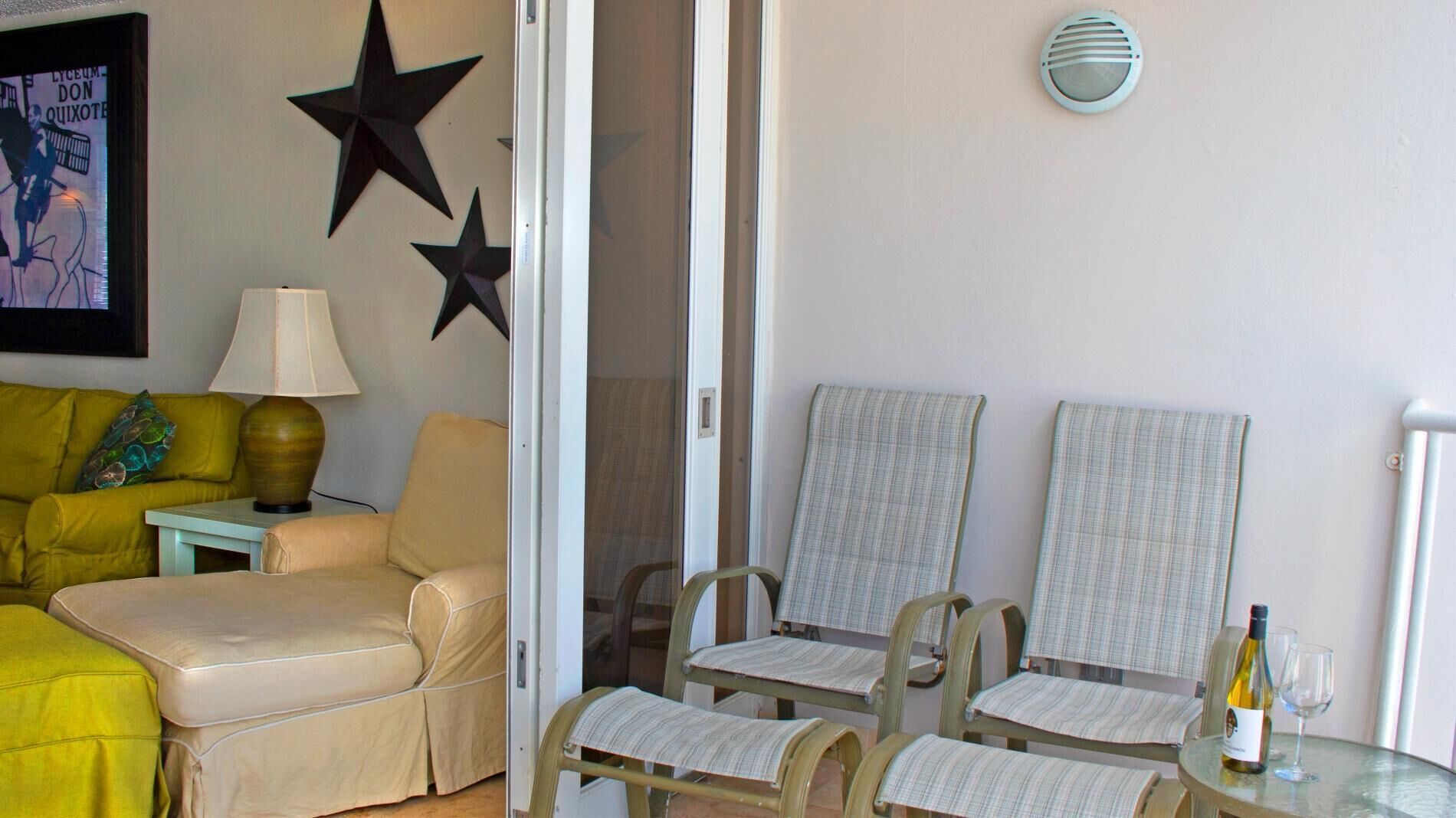Una habitación con un sofá amarillo, un sillón color crema, dos sillas a rayas, una mesa de vidrio con una botella y vasos, un adorno con forma de estrella, una lámpara y un cuadro en la pared.