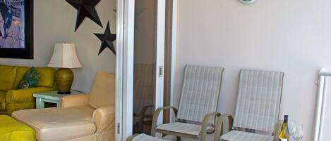 Una habitación con un sofá amarillo, un sillón color crema, dos sillas a rayas, una mesa de vidrio con una botella y vasos, un adorno con forma de estrella, una lámpara y un cuadro en la pared.