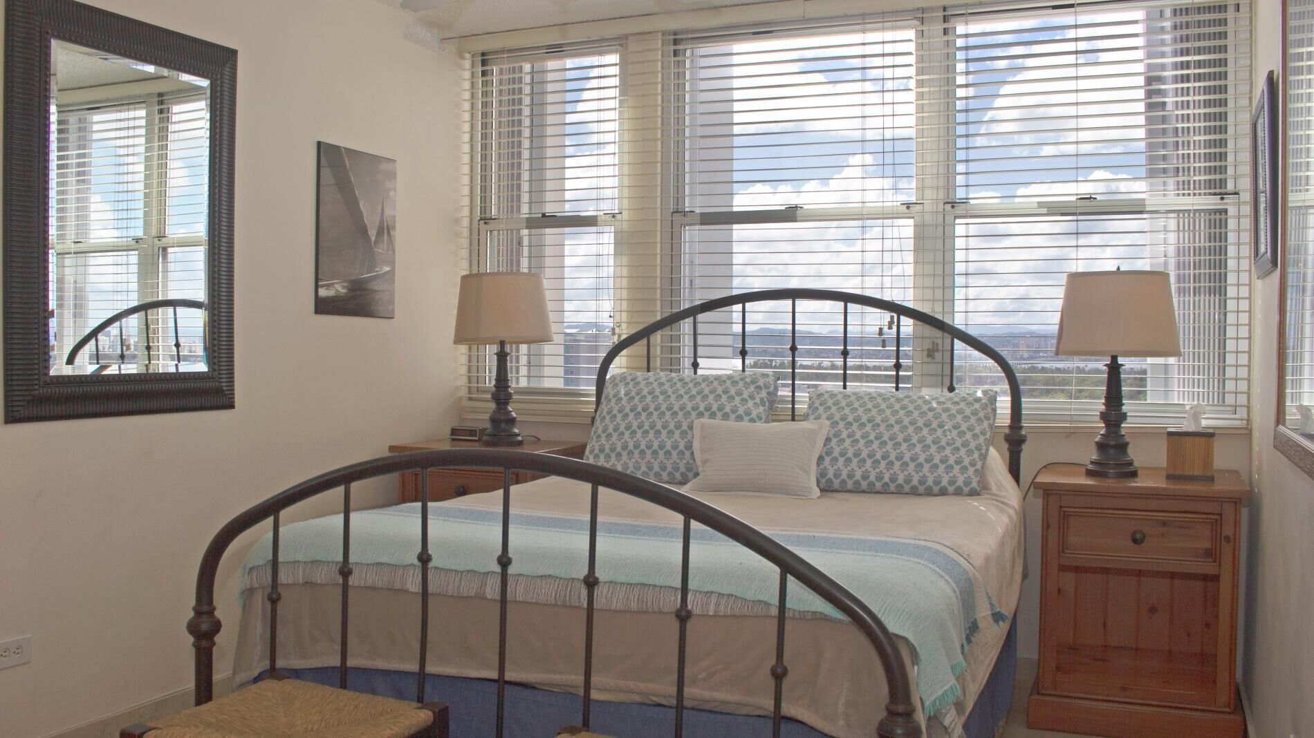 A bedroom with a metal bed frame, two bedside tables with lamps, a large window with blinds, a mirror, and a framed picture on the wall.