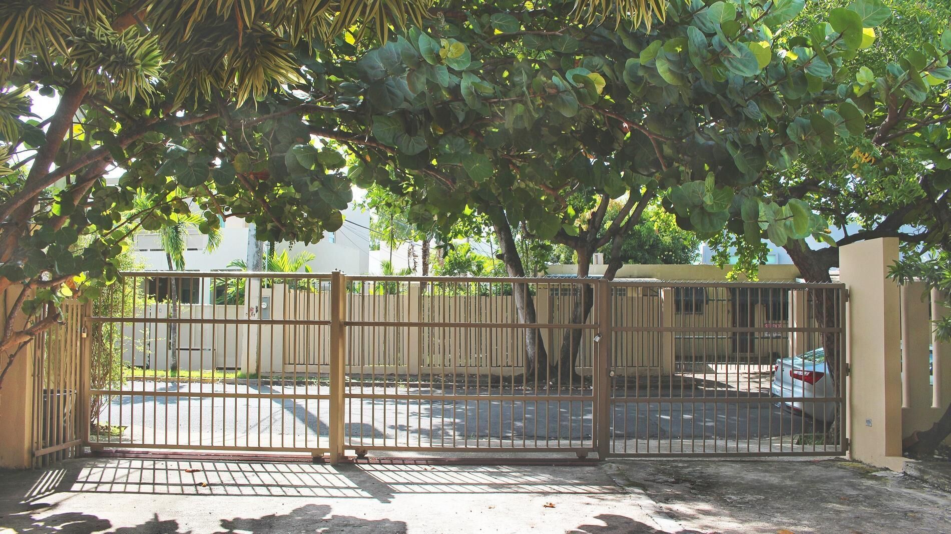 A gated parking area with a paved surface, surrounded by trees and a building in the background.