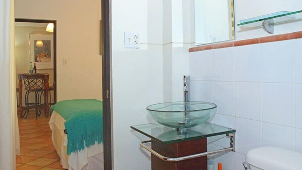 A bathroom with a glass bowl sink, a toilet, and a mirror above the sink. There is a door leading to a bedroom with a bed and a table with chairs. - Serena Baja (San Juan)