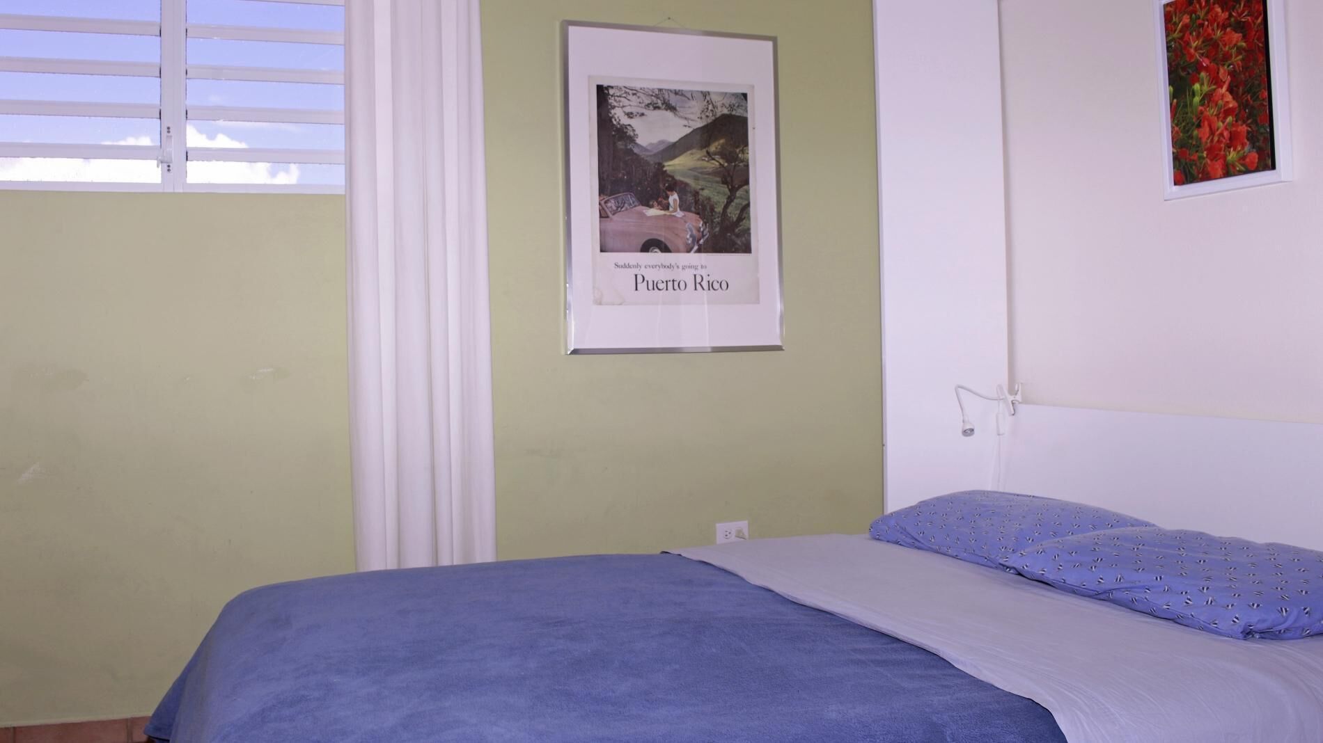 Un dormitorio con cama, una ventana con persianas blancas, un cartel en la pared y una lámpara fijada a la pared.