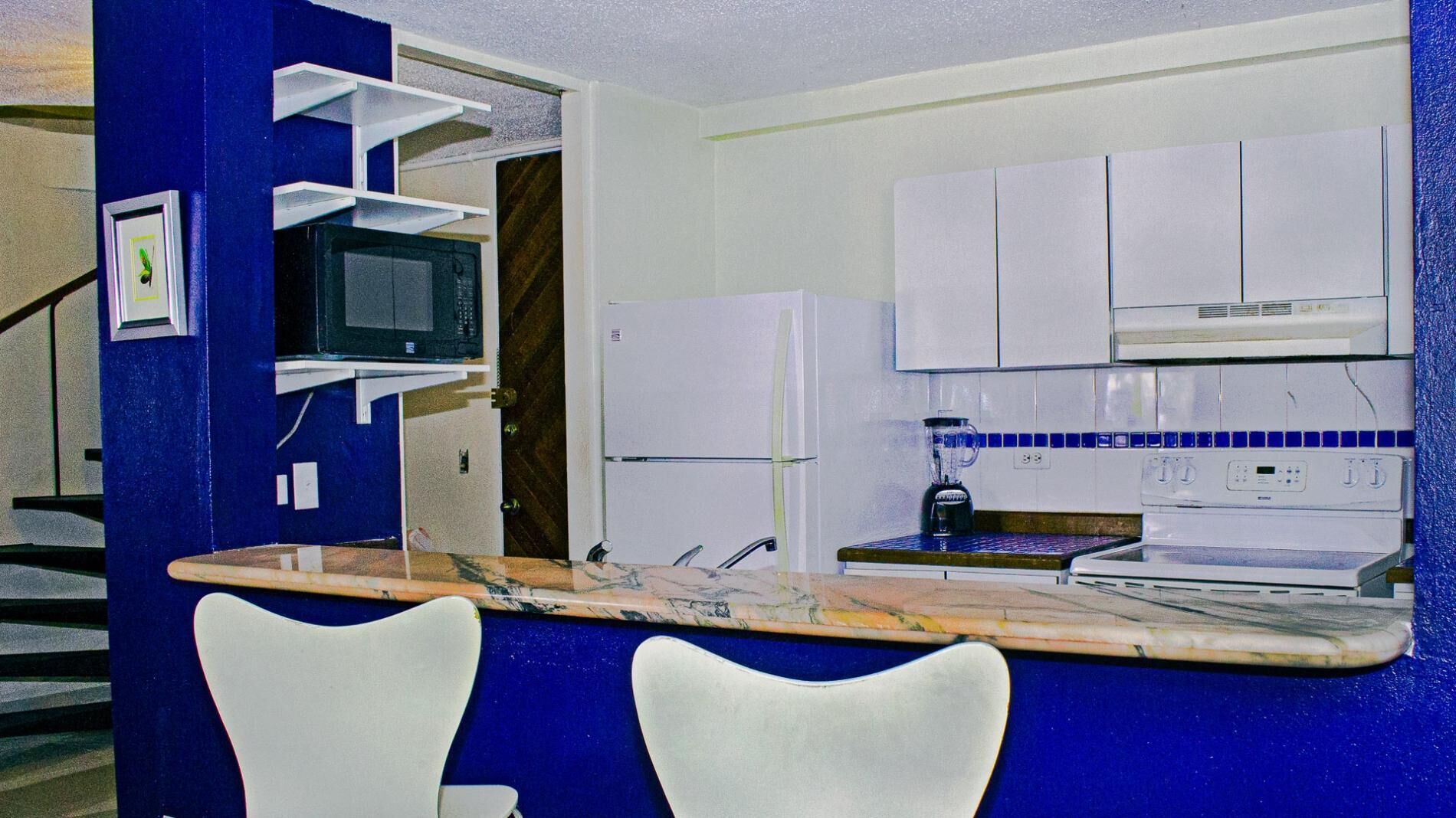 A kitchen with white cabinets, a microwave, a refrigerator, and a sink. There are two white chairs in front of the counter.