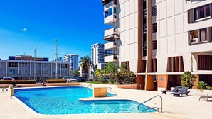 Une piscine à l’eau d’un bleu limpide, entourée d’un bâtiment blanc doté de balcons, et agrémentée de quelques palmiers.