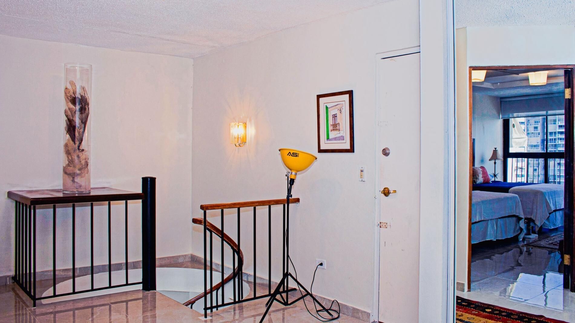 Un couloir avec un escalier, un vase en verre, un tableau encadré et une porte donnant sur une chambre avec vue sur les immebles.