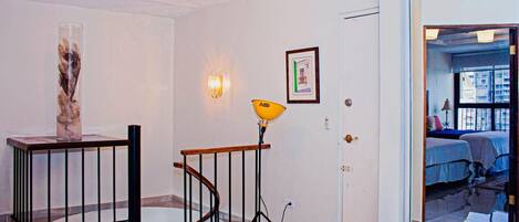 Un couloir avec un escalier, un vase en verre, un tableau encadré et une porte donnant sur une chambre avec vue sur les immebles.