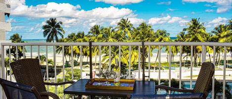 A balcony with wicker chairs and a table, overlooking a pool and palm trees.