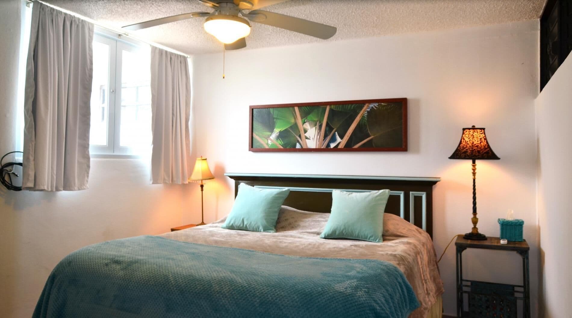 A bedroom with a bed, a ceiling fan, a window with curtains, a wall-mounted painting, and a bedside table with a lamp.