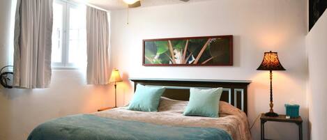 A bedroom with a bed, a ceiling fan, a window with curtains, a wall-mounted painting, and a bedside table with a lamp.