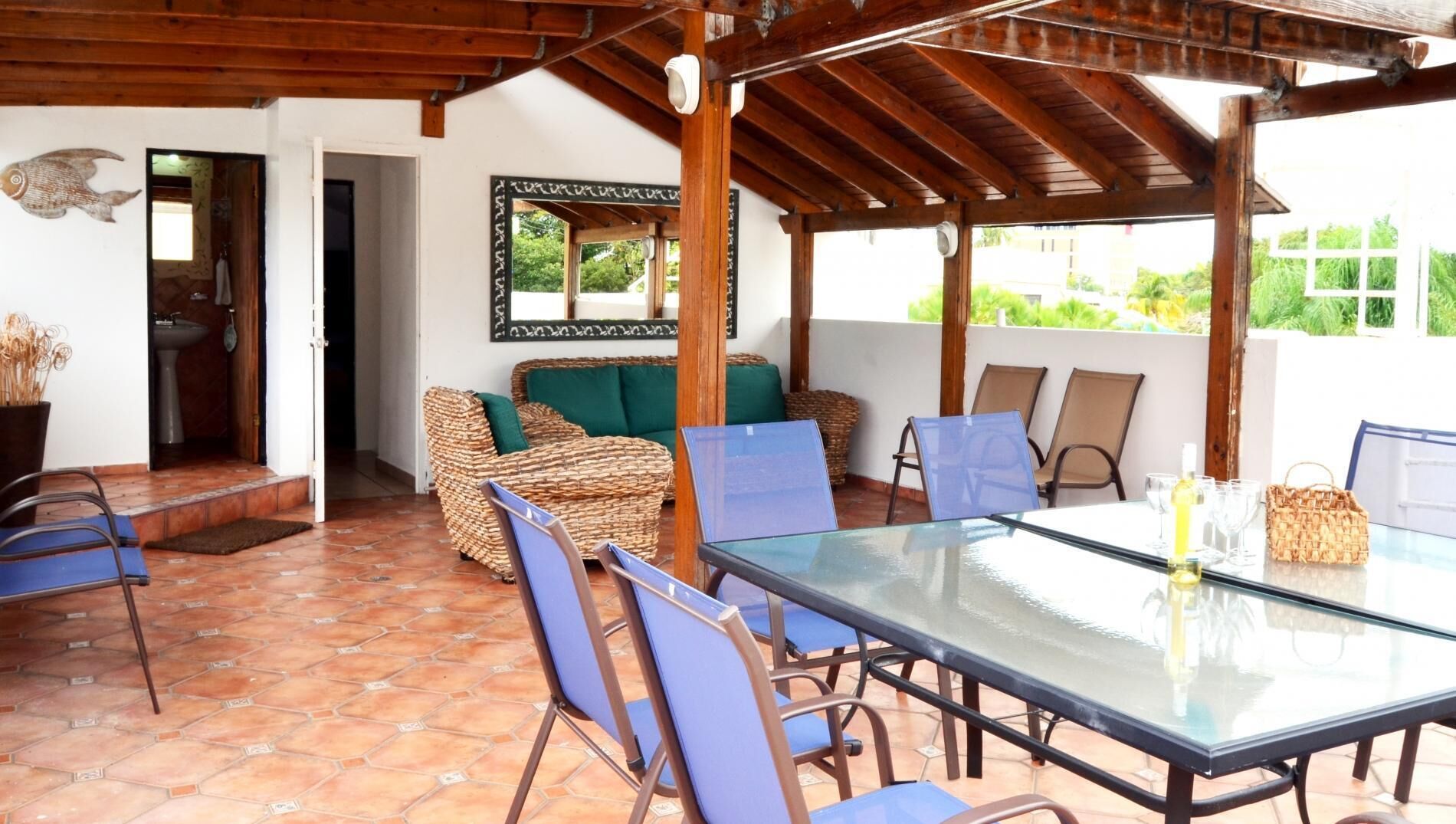A covered patio with a glass table, wicker furniture, and a view of greenery.