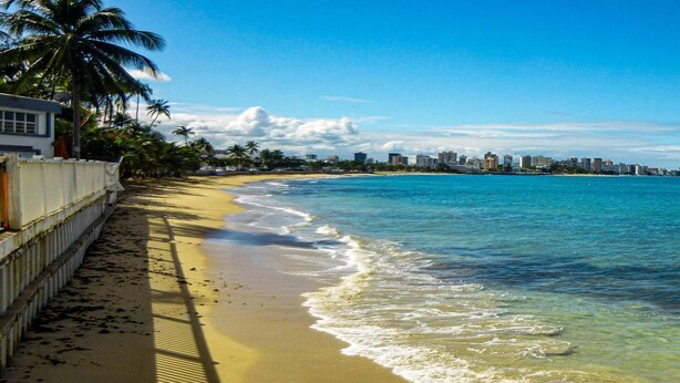 A beach with clear blue water, palm trees, and a city skyline in the background. - Casa Almendro (San Juan)
